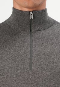 Primo piano di una persona che indossa un maglione lavorato a maglia grigio con un alto colletto a coste e una mezza zip sul davanti.