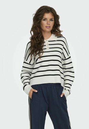 Pull à rayures blanches et noires avec un col, des boutons décoratifs et une coupe décontractée, porté avec un pantalon marine présentant des rayures latérales.