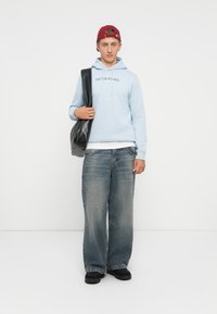 Hellblauer Hoodie mit dem Text "JACK & JONES", kombiniert mit lockeren blauen Jeans und schwarzen Schuhen; Model hält eine schwarze Tasche.