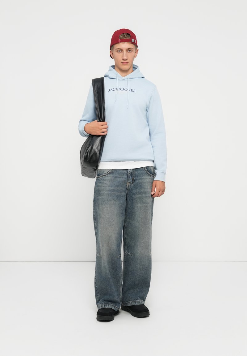 Hellblauer Hoodie mit dem Text "JACK & JONES", kombiniert mit lockeren blauen Jeans und schwarzen Schuhen; Model hält eine schwarze Tasche.