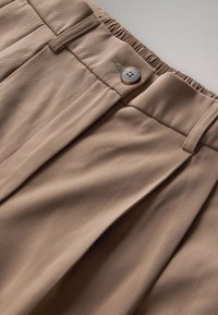 Beigenväriset korkeavyötäröiset housut, joissa laskokset, joustava vyötärönauha, vyölenkit ja nelireikäinen nappikiinnitys.