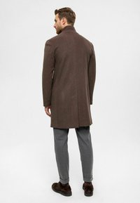 Manteau en laine brun avec un col structuré, des manches longues et une coupe droite, associé à un pantalon gris clair et des chaussures foncées.