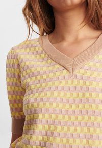 Kvinde iført en pastelrosa og gulternet kortærmet strikbluse med en beige V-hals krave.