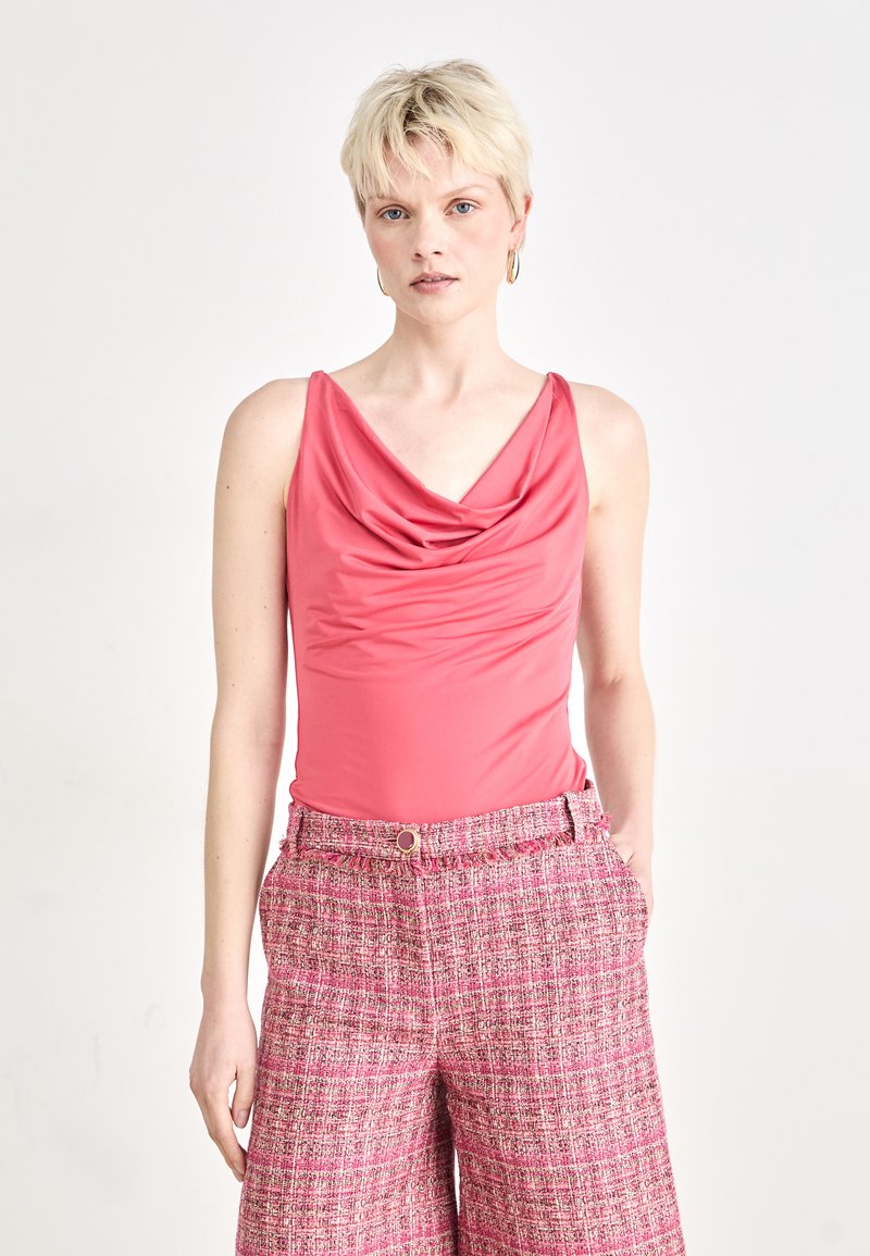 Ärmelloser Bodysuit mit Kapuze aus einfarbigem Korallstoff, kombiniert mit hochgeschnittenen, strukturierten Shorts aus pinkem Tweed, die mit einem Knopf versehen sind.