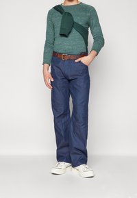 Grön texturerad långärmad skjorta med en mörkgrön tröja slängd över axlarna, marinblå byxor och vita canvas sneakers. Brun bälte.