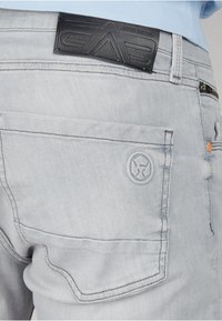 Jeans in denim grigio chiaro con una toppa in pelle in vita, caratteristiche cuciture a contrasto e un logo in rilievo sulla tasca posteriore.