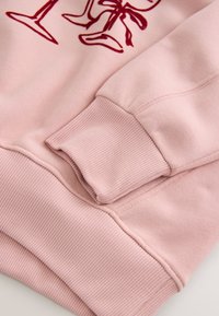 Ružová mikina s rebrovanými manžetami a lemom, ktorá má hladkú textúru a grafický potlač v bordovej farbe. Dizajn s voľným strihom.