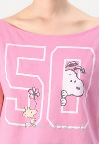 Růžové bavlněné tričko s velkým bílým grafickým číslem "50" a kreslenými postavičkami Snoopyho a Woodstocka, kteří drží květinu, s mírně posunutými rameny.