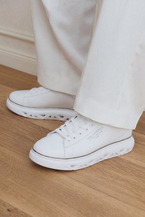 Λευκά χαμηλά sneakers με μοναδικές σόλες με κοψίματα, φορεμένα με ανοιχτόχρωμο παντελόνι σε ξύλινο πάτωμα.