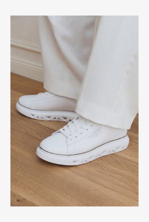 Λευκά χαμηλά sneakers με μοναδικές σόλες με κοψίματα, φορεμένα με ανοιχτόχρωμο παντελόνι σε ξύλινο πάτωμα.