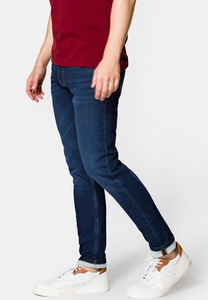 Ciemnoniebieskie, dopasowane dżinsy wąskie noszone z białymi trampkami z brązowymi akcentami; materiał wydaje się gładki z lekkim streczem. Wywinięte nogawki.