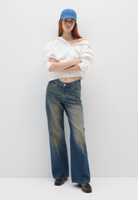 Sudadera blanca con cremallera, gorra de denim azul claro, jeans de pierna ancha en un azul desgastado, textura suave, zapatos negros planos, de pie contra un fondo liso.