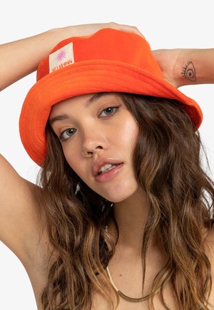 Cappello a secchiello arancione realizzato in tessuto morbido, con una visiera cucita e un'etichetta quadrata con un logo a forma di sole. Design semplice con finitura testurizzata.