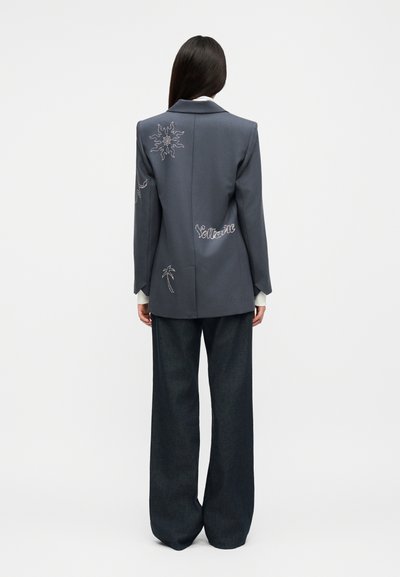 Γυναίκα με μακριά σκουρόχρωμα μαλλιά, που φοράει γκρι σακάκι με κέντημα ήλιου, φοινικόδεντρου και τη λέξη "Solitaire", συνδυασμένο με φαρδιά σκούρα παντελόνια, που στέκεται γυρισμένη προς τα πίσω.