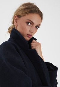 Marineblauer Wollmantel mit hohem Kragen, der ein strukturiertes Design und eine weiche Textur aufweist. Das Modell hält den Kragen und präsentiert die satte Farbe des Stoffes.