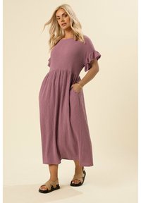 Abito maxi in misto lino color malva con maniche corte a volant, tessuto strutturato e due tasche laterali. Indossato con sandali neri e cinturini beige.