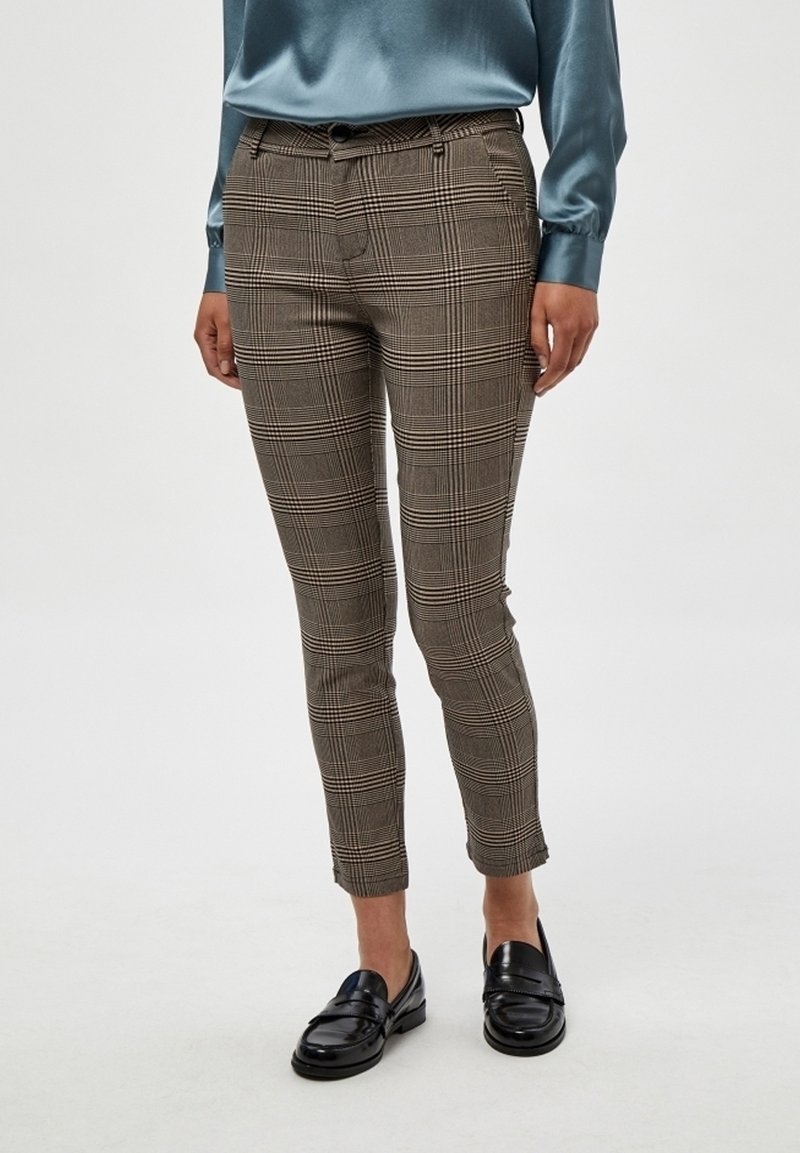 Personne portant un pantalon slim marron à carreaux, un chemisier en satin bleu sarcelle à manches longues et des mocassins en cuir noirs, debout sur fond blanc.