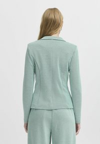 Mintgrön blazer med en texturerad tyg, skräddarsydd design, spetsiga slag och strukturerade axlar, visad bakifrån.