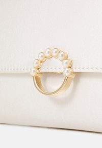 Kremowa satynowa kopertówka z okrągłym złotym uchwytem ozdobionym sześcioma małymi perłami, o gładkiej teksturze i starannym przeszyciu.