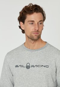 Sudadera gris hecha de una tela suave, con un escote a la caja y el texto "SAIL RACING" bordado en azul oscuro en el pecho.