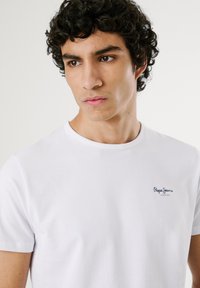 Junger Mann mit dunklen lockigen Haaren, der ein weißes Pepe Jeans London T-Shirt trägt und nach unten sowie leicht zur Seite blickt, vor einem schlichten Hintergrund.