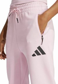 Ljusrosa mjukisbyxor med elastisk midja, justerbar snörning, sidoficka och svart adidas-logotyp på vänstra benet. Slät textur.