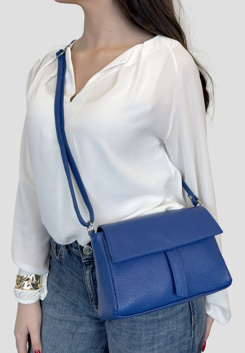Vrouw draagt een witte blouse en spijkerbroek, met een blauwe leren crossbodytas met textuur en verstelbare riem over haar schouder.
