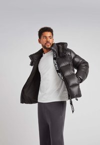 Giacca a vento nera con cappuccio, caratterizzata da una finitura lucida, design trapuntato e una nastro con logo bianco a contrasto. Abbinata a una camicia bianca.