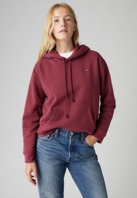 Maroon hoodie with a front pocket and adjustable drawstring hood, paired with blue denim jeans. Ribbed cuffs and seamless design.