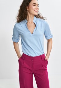 Jasnoniebieska bluzka z krótkim rękawem i dekoltem w serek, z podwiniętymi mankietami, w zestawieniu z wysokim stanem fioletowymi spodniami z podwójnymi guzikami i bocznymi kieszeniami.