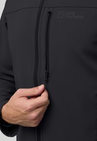 Schwarze Softshelljacke aus strukturiertem Material, mit vorne befindlichem Reißverschluss und einem Logo auf der Brust. Hand ist zu sehen, die den Reißverschluss zieht.