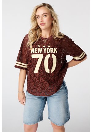 Jonge vrouw met lang blond haar, gekleed in een doorschijnend bordeauxrood kanten New York 70-shirt en lichte denim shorts, staand met één hand op haar heup.