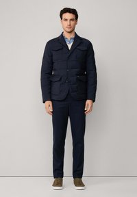 Marineblauwe gewatteerde blazer met meerdere voorkruiden, gedragen over een wit trui en een blauw gestreept shirt, gecombineerd met gestreepte marineblauwe broek en olijfgroene sneakers.