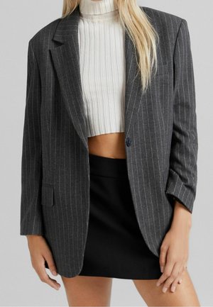 Vrouw draagt een donkergrijze blazer met krijtstreep over een witte geribbelde cropped top en een zwarte rok, poserend met ontspannen handen langs haar zij.