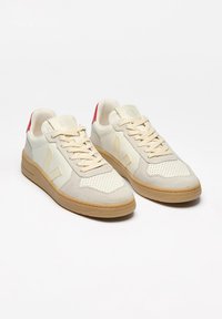 White and light beige sneakers with suede accents, perforated toe box, and red heel tab. Features beige laces and gum rubber soles.