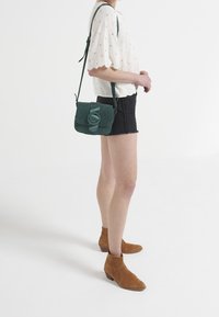Teal suede crossbody bag with a flap closure and circular accent, accompanied by a short-sleeve white top and black denim shorts.