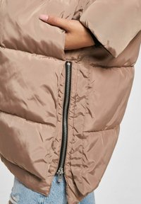 Hånd i lommen på en beige polstret jakke med stor sølvglidelås, båret over lys blå ripped jeans.