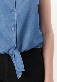 Camicia in denim blu con nodo legato all'orlo, dettagli a bottoni e maniche corte. Indossata con pantaloni neri, presenta uno stile casual.
