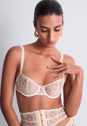Femme aux cheveux lisses porte un soutien-gorge en dentelle blanche transparente et une lingerie assortie taille haute, touchant sa poitrine et montrant une bague en or et des boucles d'oreilles.