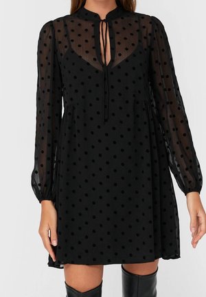 Mujer con un vestido negro transparente de lunares con mangas largas, detalle de lazo en el cuello y botas negras hasta la rodilla.
