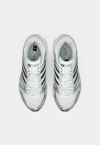 Bijele atletske tenisice s mrežastim gornjim dijelom, crnim prugama i srebrnim detaljima. Imaju vezanje na vezice i podstavljene potplate.