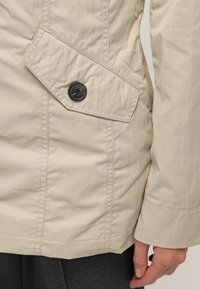 Chaqueta de color beige claro hecha de tejido arrugado, que presenta un bolsillo con solapa diagonal con un botón oscuro. Los detalles de las costuras son visibles a lo largo de los lados.