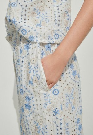 Hand in pocket of white outfit with blue and beige floral embroidery and geometric cutout patterns on fabric.
