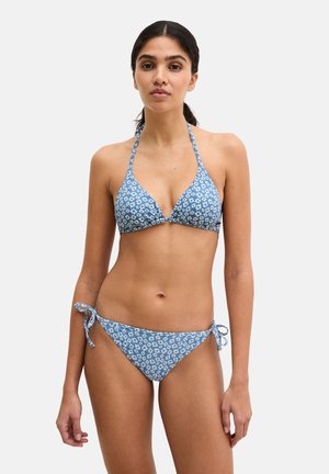 Woman standing wearing a blue halter-neck bikini with white floral print and tie-side bottoms against a plain light background.