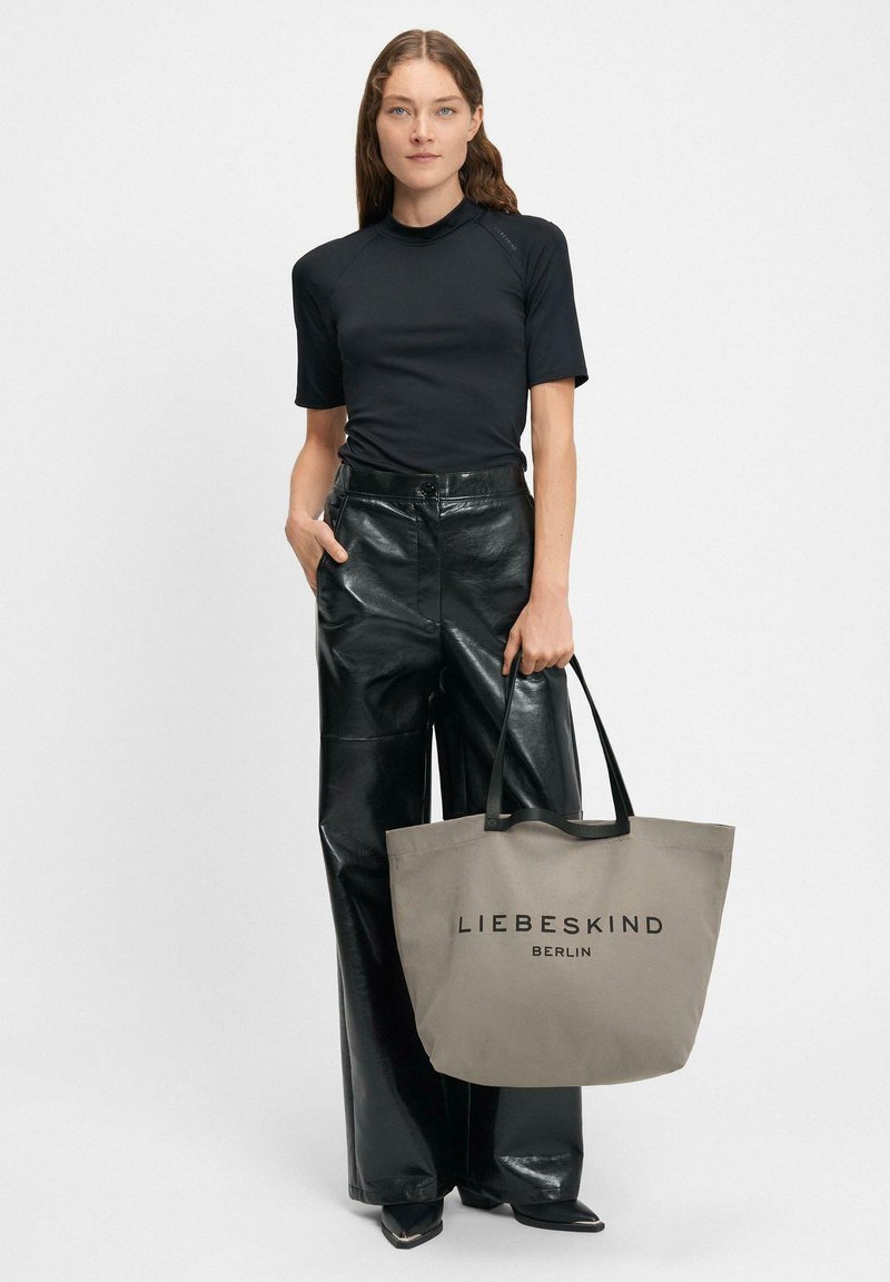 Kobieta w czarnej bluzce z krótkim rękawem i błyszczących czarnych spodniach, trzymająca dużą szaro-skalną torbę z logo "LIEBESKIND BERLIN".