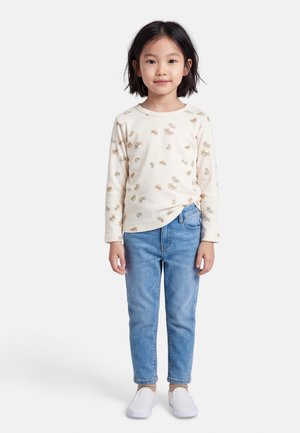 Nuori tyttö, jolla on lyhyet mustat hiukset, beige pitkähihaispaita, jossa on rapukuvio, vaaleansiniset farkut, beiget sukat ja valkoiset slip-on-kengät.