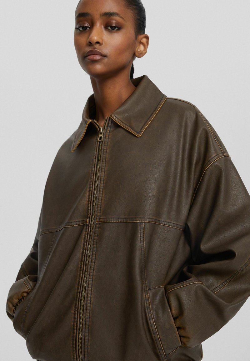 Joven mujer de piel oscura que lleva una chaqueta de cuero marrón oscura holgada con cremallera frontal y las manos en los bolsillos, fondo neutro.