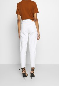 Kvinna i högmidjade vita jeans, bränd orange t-shirt med kort ärm och svarta remsandaler med hög klack, som står mot en enfärgad bakgrund.