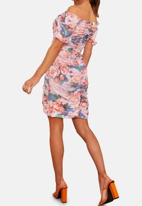 Nainen, joka käyttää vaaleanpunaista kukallista olkapäätöntä rypytettyä mekkoa ja oransseja korkeakorkoisia sandaaleja, seisoo yhdellä jalalla hieman eteenpäin.