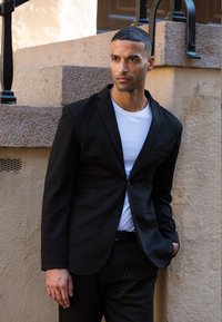 Sort blazer med en enkelt knap og lomme, iført over en hvid t-shirt. Suiten er figursyet og står foran en struktureret beige væg.
